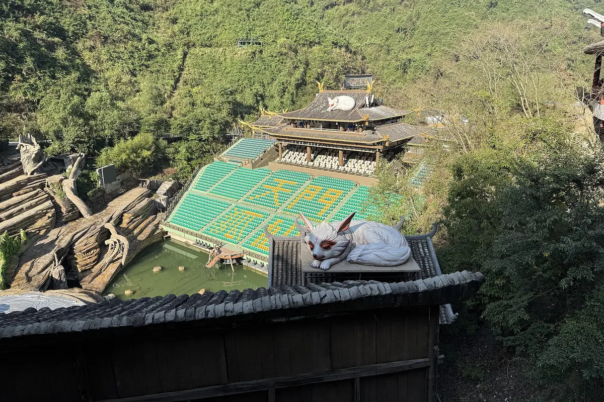 The Tianmen Fox Fairy theatre by day: tiers of green seats banked up a wooded valley slope, a pagoda-roofed building above them, and a white fox statue on a roof in the foreground